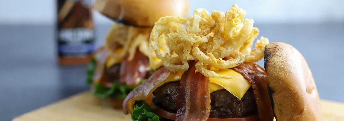 burger open to reveal fried onions and bacon with another burger and a bottle of seasoning in the background