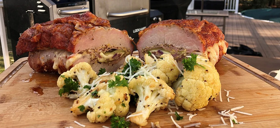 pork loin cut in half with garnished and cooked cauliflower on wood board with pit boss grill in background