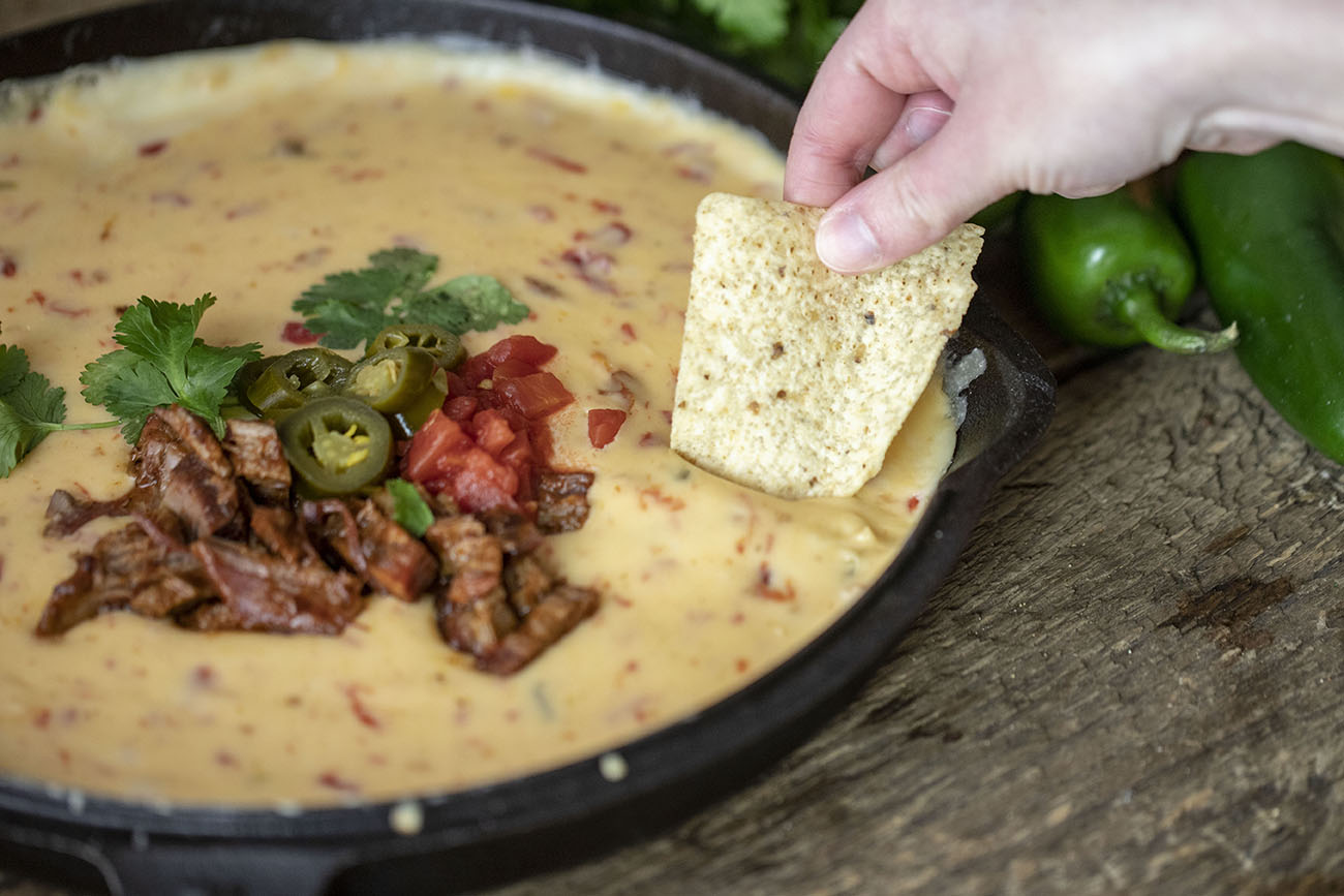 brisket queso with jalapeno and tomatoes on top next to Pit Boss Sweet Rib Rub bottle, jalapenos and lemons