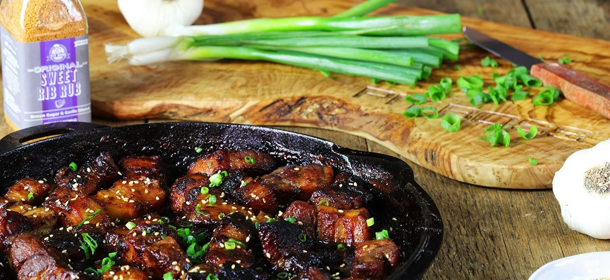 braised pork belly in cast iron skillet next to bunch of green onion on wood board and sweet rib rub