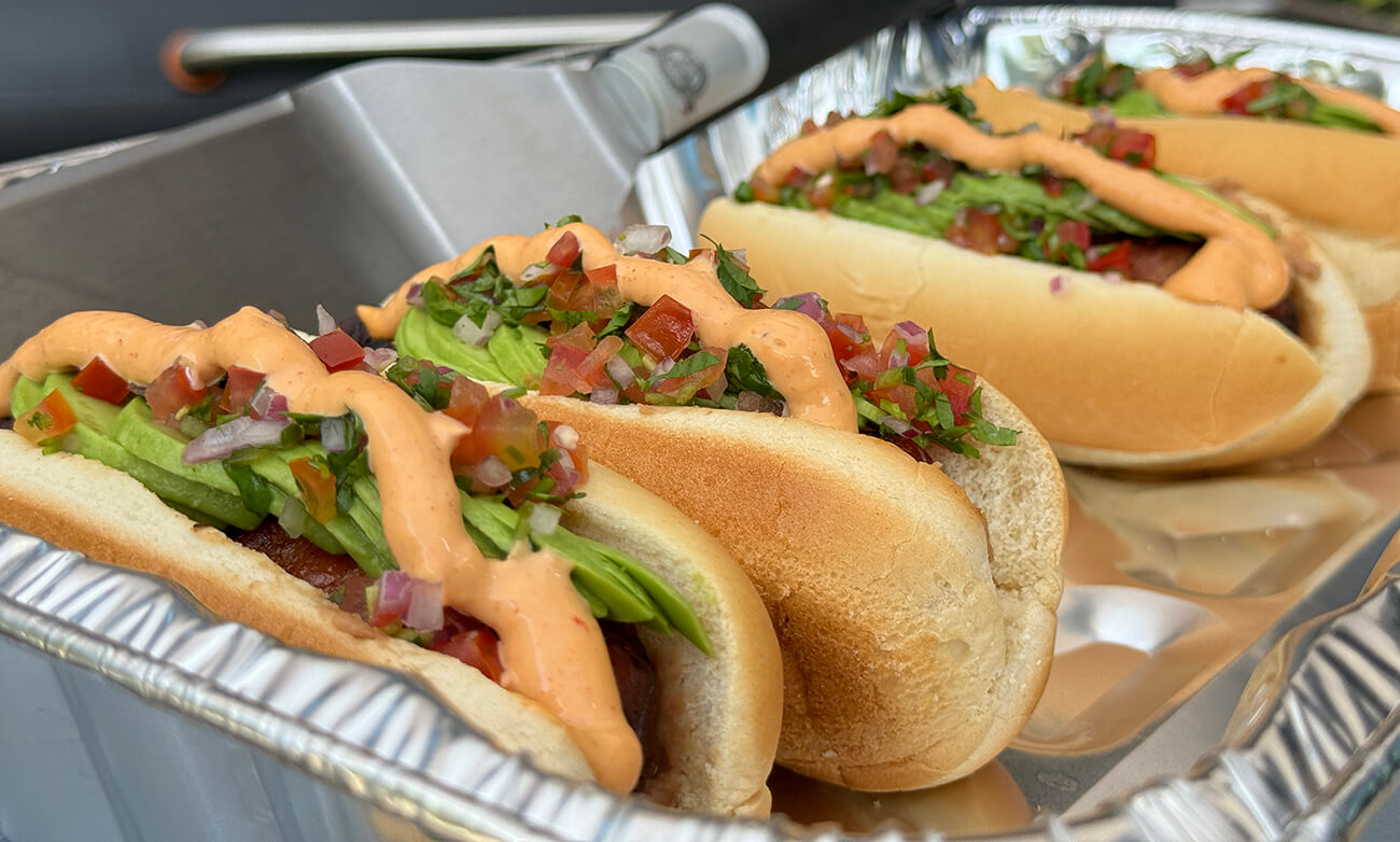 grilled sonoran hot dog cooked on pit boss wood pellet grill in aluminum pan with pico de gallo, avocado and chipotle mayo