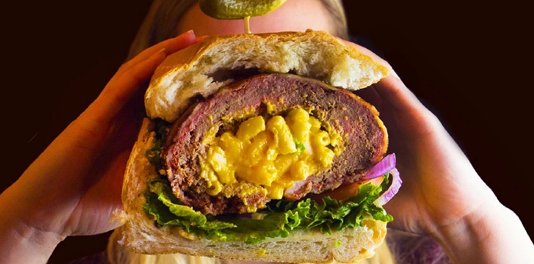two hands holding a macaroni-stuffed bacon-wrapped burger