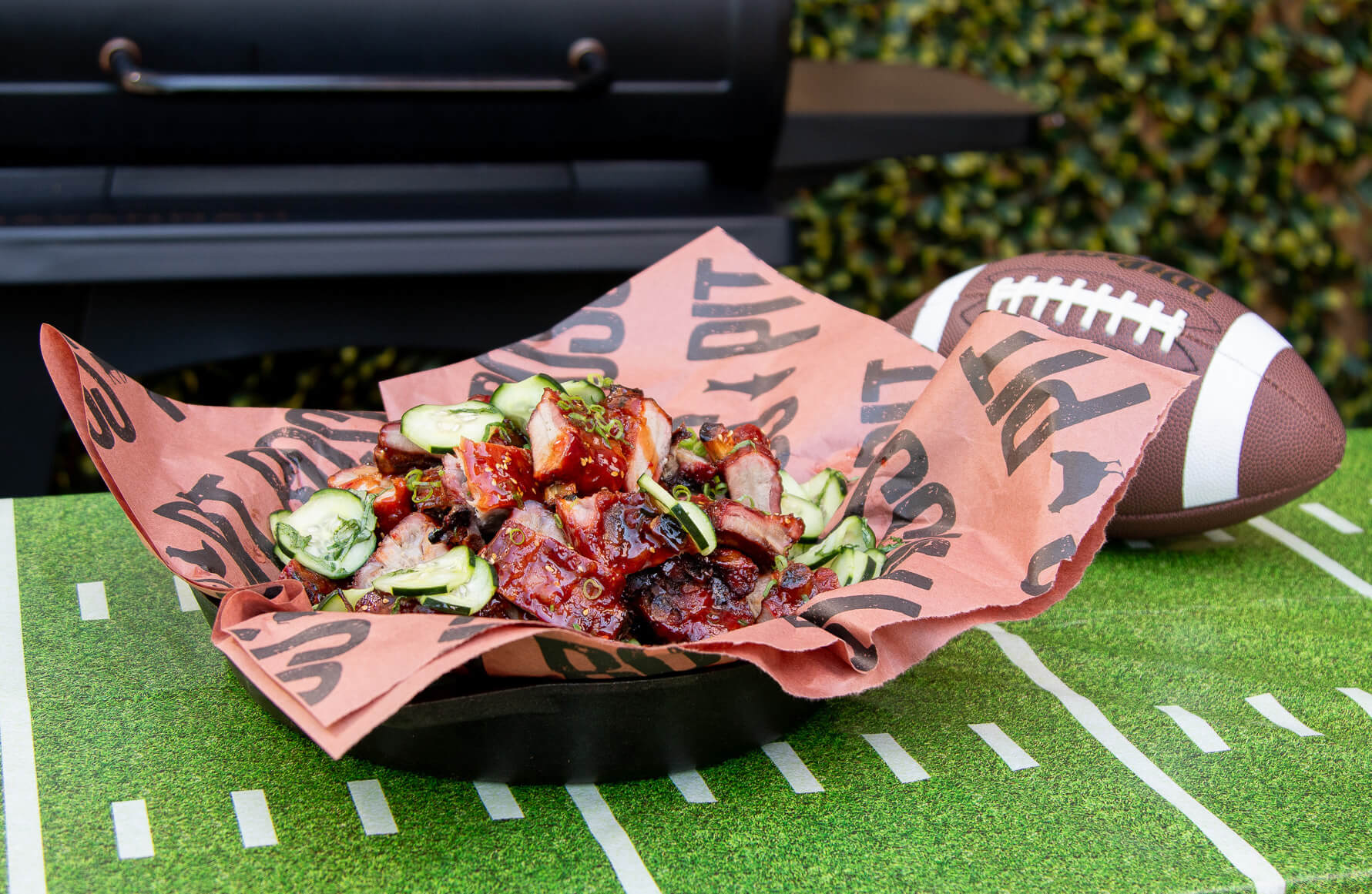 glazed smoked baby back ribs garnished with marinated cucumbers in cast iron dish in front of pit boss wood pellet grill. game day appetizer