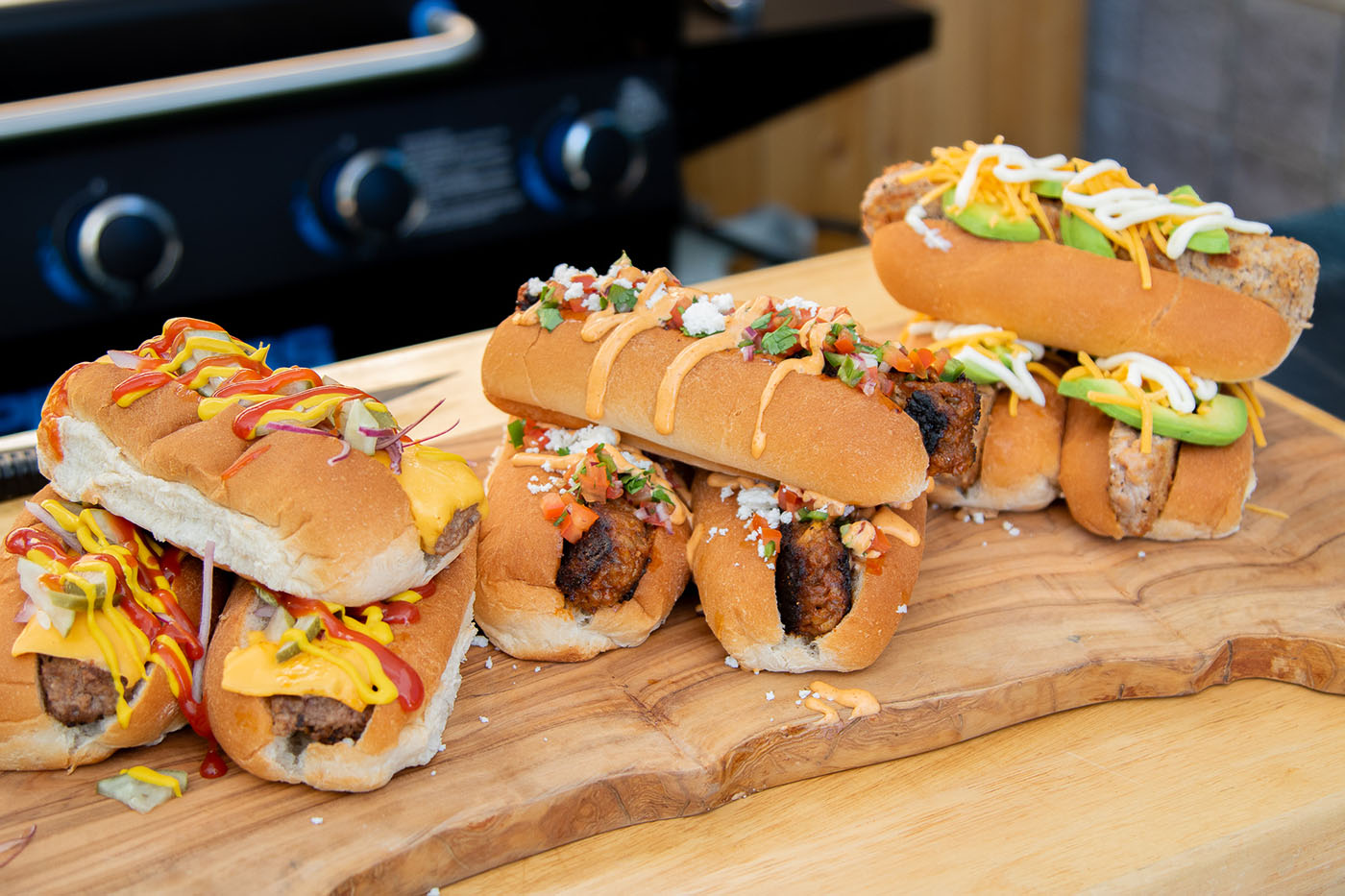 3 burger dog recipes cooked on a pit boss gas griddle and served on wooden cutting board with various toppings