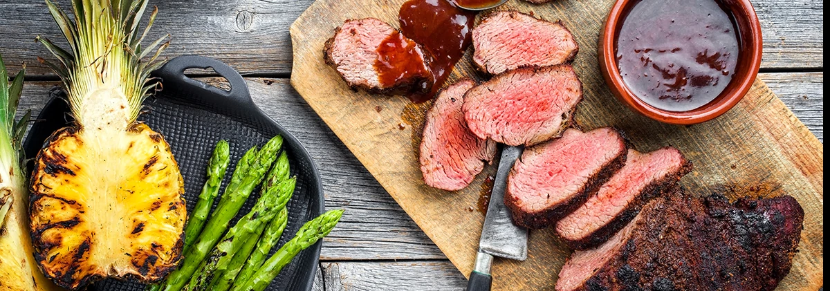 sliced steak on wood cutting board next to sauce and grilled pineapple and asparagus