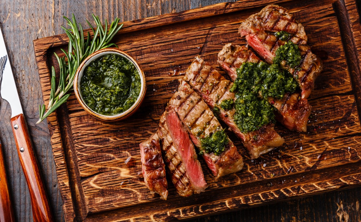 Sliced rare steak on wood board next to bowl of chimichurri sauce and herbs